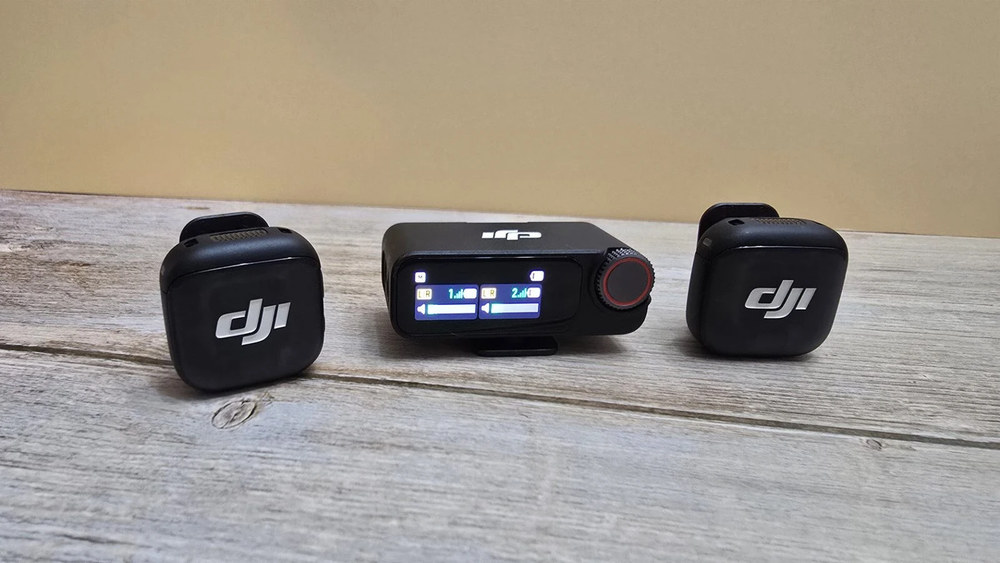Два квадратні передавачі та один прямокутний приймач DJI Mic 3 з сенсорним екраном і червоно-сірим коліщатком лежать на дерев'яному столі.