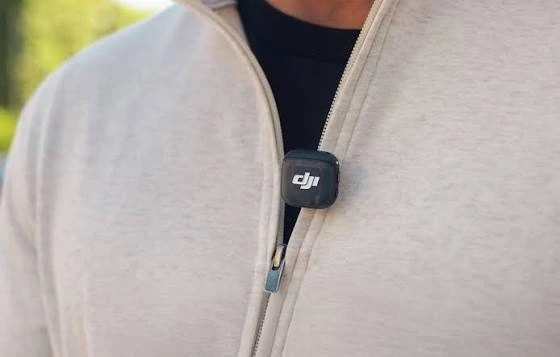 Чорний квадратний передавач DJI Mic 3 закріплений на блискавці світлої кофти користувача, на корпусі видно білий логотип бренду та зелений індикатор.
